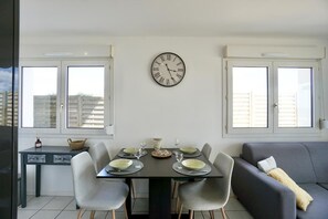 A dining area with a table set for four, a clock on the wall, and a sofa in the background. - Swimming pool with a 100-meter cable winch, located 100 meters from the beach* (Guidel)