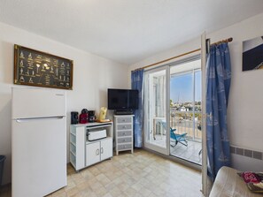 Quarto compacto com geladeira, cafeteira, televisão e vista para um porto através de uma porta de vidro deslizante.