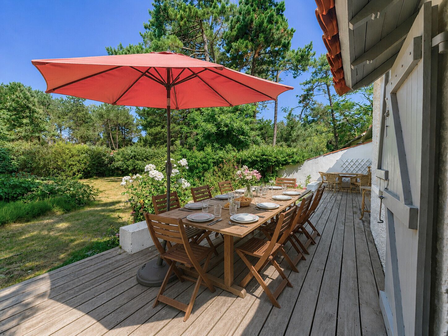An outdoor wooden deck with a dining table set for four, surrounded by chairs, and a red umbrella providing shade.