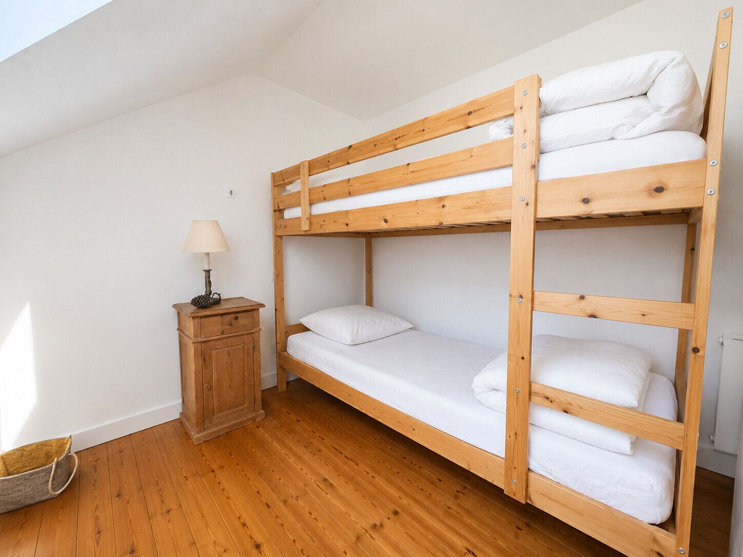 A bunk bed with white bedding, a wooden nightstand with a lamp, and a small basket on the floor.