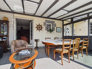 Eine sonnendurchflutete Terrasse mit einem runden Tisch und Stühlen, einem Bücherregal und einer Tür zu einem Innenbereich.