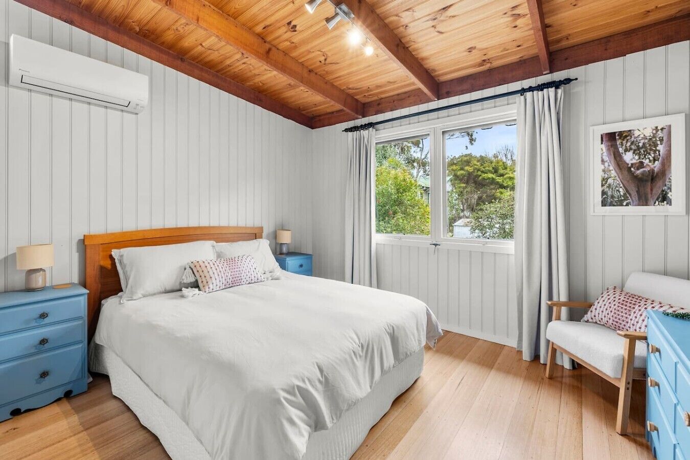 A bedroom with a bed, nightstands, a chair, a window with curtains, and a wall-mounted air conditioner.
