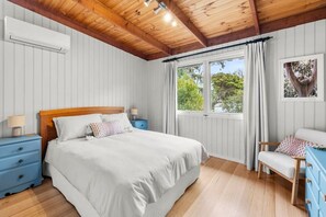 Ein Schlafzimmer mit einem Bett, Nachttischen, einem Sessel, einem Fenster mit Vorhängen und einer Wand-Klimaanlage.