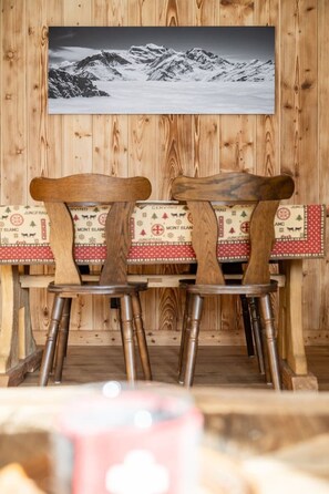 Een houten eettafel met twee stoelen, een houten wand met een lijstfoto en een tafelkleed met dessin.