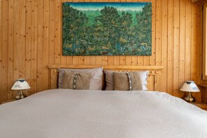 A bedroom with a wooden paneled wall, a large bed with a white bedspread, two bedside lamps, and a painting of a forest scene above the bed. - Cozy, Alpine • Terrace • Indoor parking (Val de Bagnes)