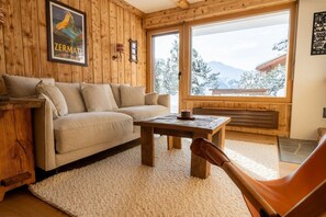 Een gezellige kamer met een bank, een salontafel en een stoel. Er is een raam met uitzicht op met sneeuw bedekte bomen en bergen.