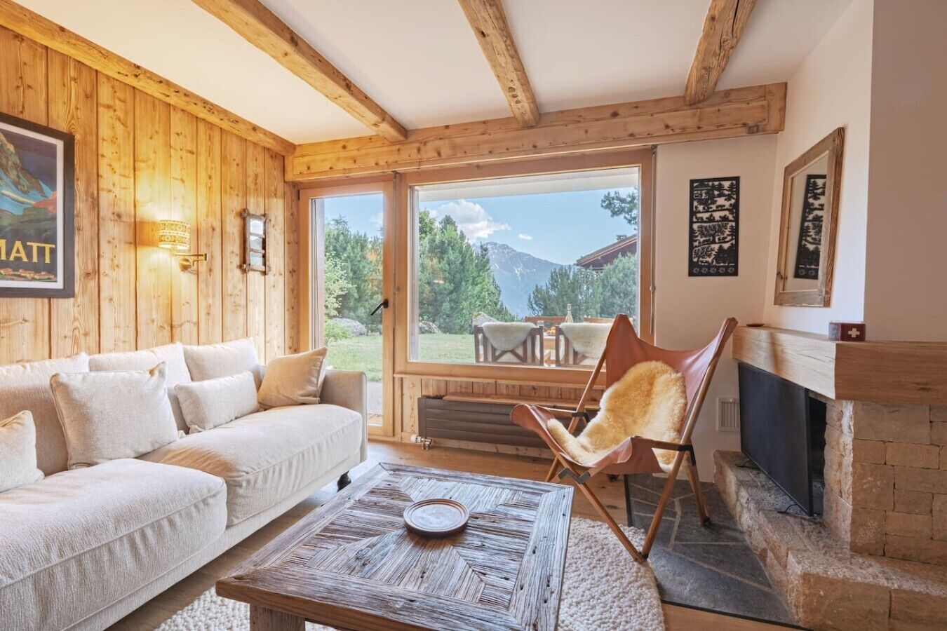 A cozy living room with a wooden ceiling, a sofa, a chair, a fireplace, and a large window offering a mountain view.