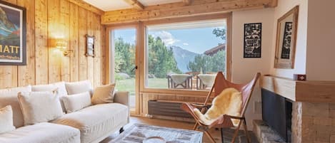 A cozy living room with a wooden ceiling, a sofa, a chair, a fireplace, and a large window offering a mountain view.
