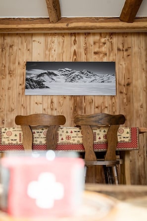 A wooden interior with a framed picture of a snowy mountain landscape on the wall, wooden chairs with patterned cushions, and a table with a pink object in the foreground. - Cozy, Alpine • Terrace • Indoor parking (Val de Bagnes)