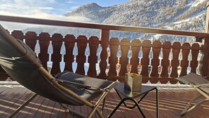 Een houten terras met uitzicht op besneeuwde bergen. Twee ligstoelen en een tafel met een kaarsje staan er.
