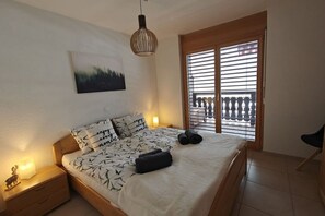 Quarto com cama, mesa de cabeceira, luminária na parede, janela com persianas e vista para a varanda.