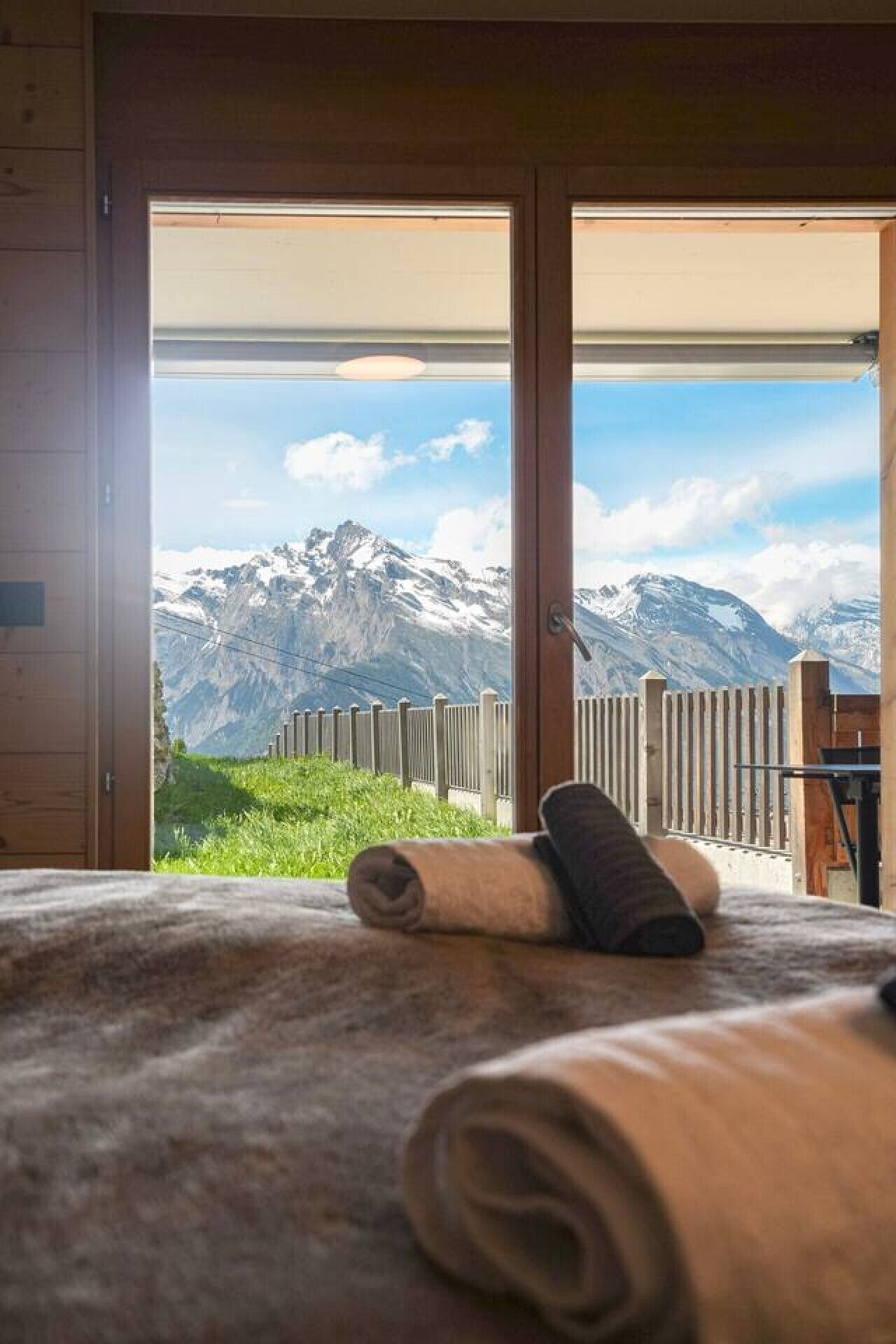 A room with a large window offering a mountain view, a wooden floor, and a rolled-up towel on the bed.