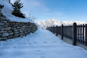 Un sendero cubierto de nieve flanqueado por un muro de piedra y una valla de madera que conduce a una cadena montañosa.