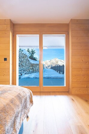 Ein Zimmer mit Holzboden, einem Bett mit Felldecke und einem großen Schiebeglasfenster, das auf eine verschneite Landschaft mit Steinmauer und Holzzaun blickt.