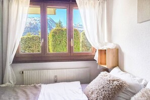 A bedroom with a view of mountains through a window, a wooden bedside table, and a fluffy pillow. - Cozy & Bright • 1 Bedroom • Ovronnaz (Leytron)