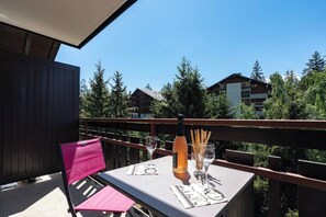 Een balkon met een tafel gedekt voor twee personen, een fles en glazen, met uitzicht op een bebost gebied met gebouwen op de achtergrond.