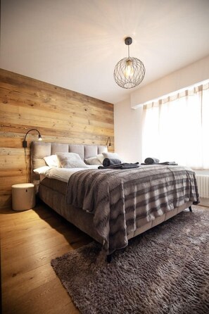 Een slaapkamer met een houten wand, een bed met een grijze deken, een ronde lamp en een raam met gordijnen.