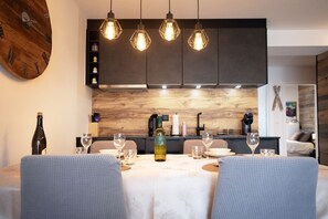 Een eetgedeelte met een houten klok, hanglampen, een wijnfles en een tafel gedekt voor vier personen.