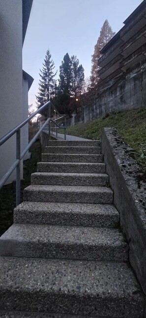 A set of concrete stairs leading up to a building entrance, with a metal handrail on the left and a retaining wall on the right. - Modern Apartment with Mezzanine – Stunning Village View (Zermatt)