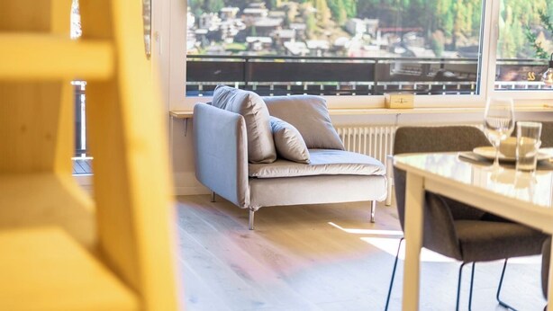 Een moderne woonkamer met een bank, een eettafel en uitzicht op een bebost gebied door grote ramen.