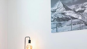 Een bed met een witte deken en een plaid met een wegenkaart ontwerp, een bedlamp, een ingelijst berglandschap en een houten nachtkastje.