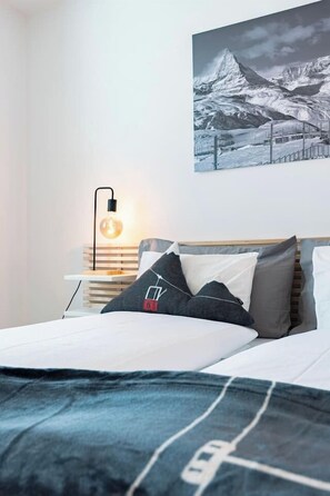 Ein Bett mit weißer Bettdecke und einer Decke mit einer Straßenkarten-Musterung, eine Nachttischlampe, ein gerahmtes Bild einer Berglandschaft und ein Nachttisch aus Holz.