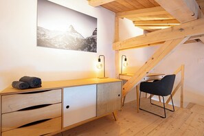 A modern bedroom with a wooden desk, a chair, a bed, and a mountain landscape painting. - Modern Apartment with Mezzanine – Stunning Village View (Zermatt)