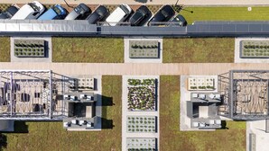 Vista aérea de un estacionamiento con múltiples filas de coches aparcados, un área de césped verde y un edificio con jardín en la azotea.