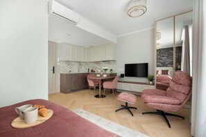 A modern bedroom with a bed, a pink armchair, a TV, and a kitchen area with a marble countertop.