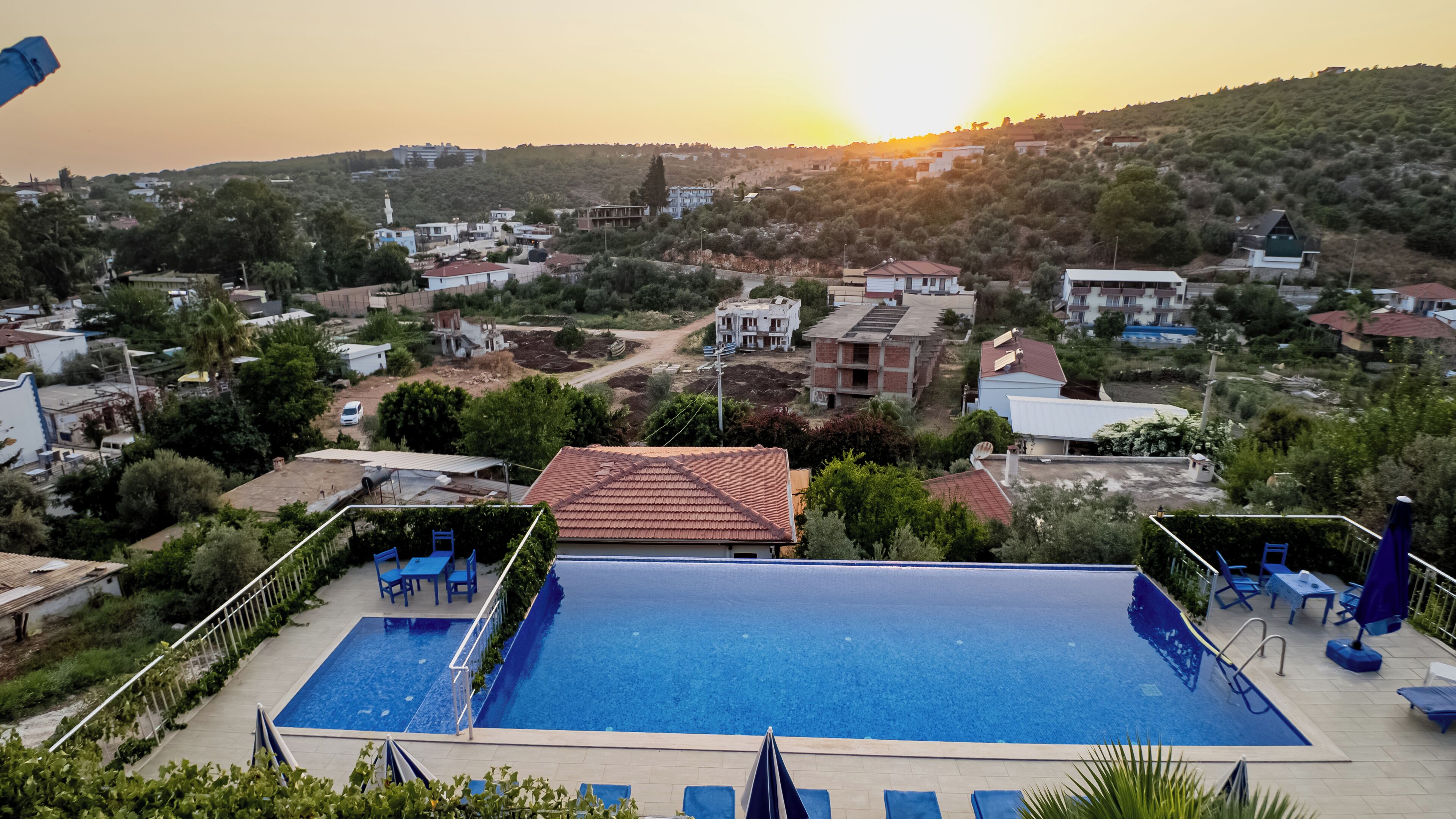 A swimming pool with lounge chairs and a view of a residential area during sunset.