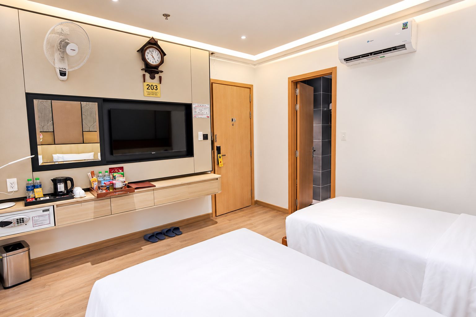 Habitación de hotel con cama, mesita de noche, televisor, reloj y una pequeña zona de kitchenette.