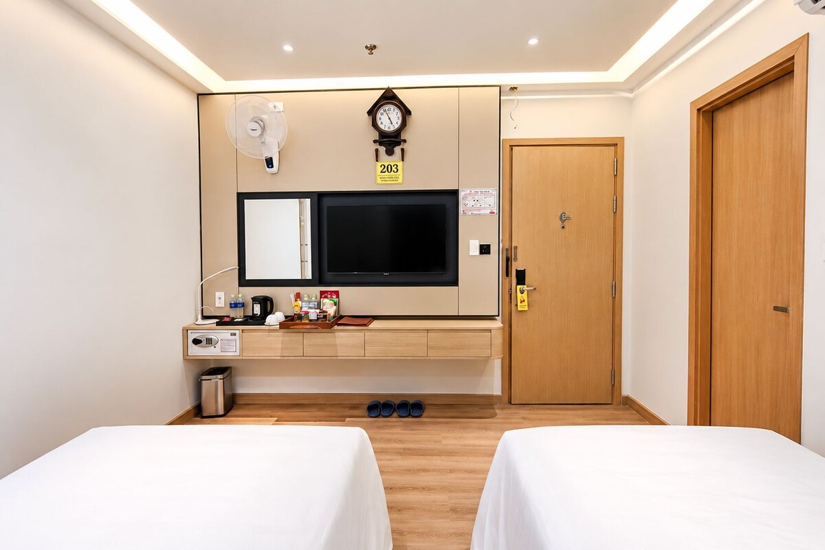 A hotel room with two beds, a TV, a clock, a fan, a small table with items, and a door.