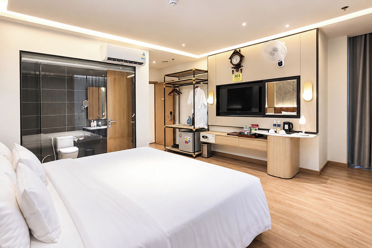 A modern hotel room with a large bed, a built-in TV, a wardrobe, a vanity with a mirror, and a bathroom with a glass shower enclosure.