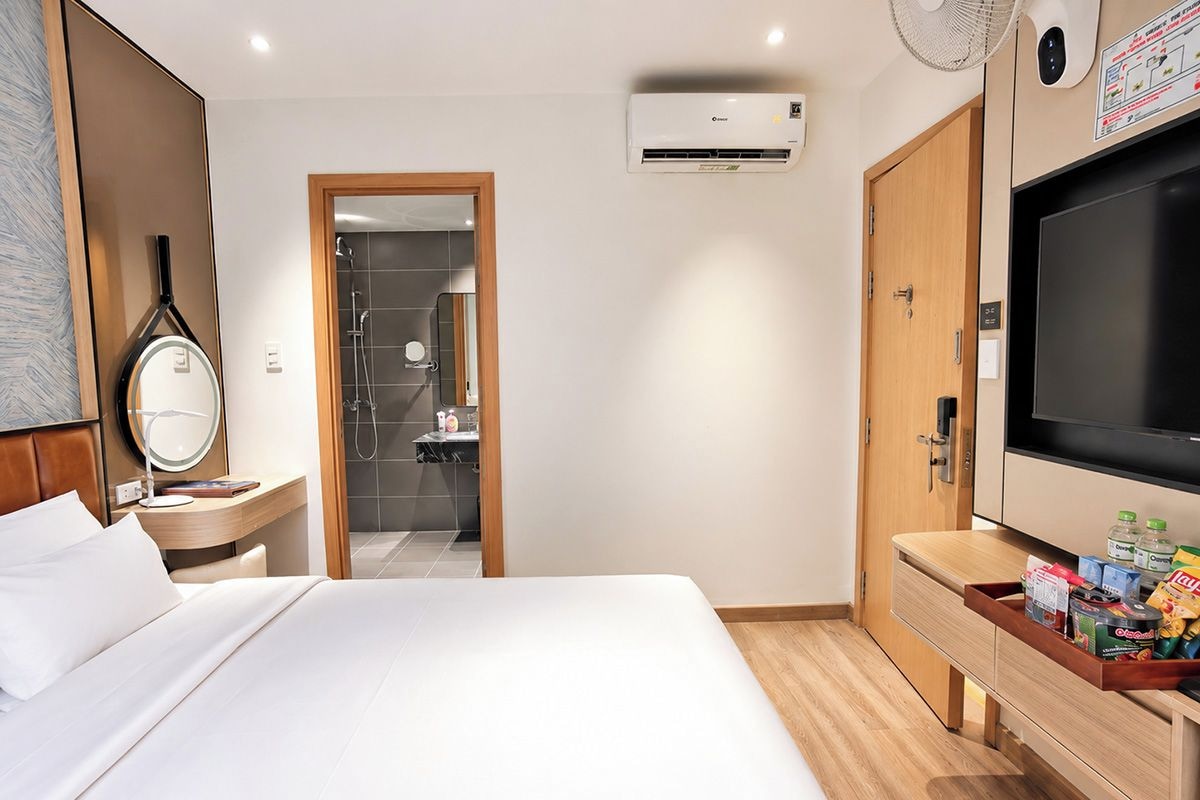 A hotel room with a bed, bedside table, mirror, TV, wardrobe, and a small kitchenette area.