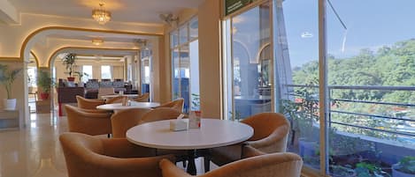 Een hotellobby met ronde tafels en stoelen, grote ramen en uitzicht op groen.