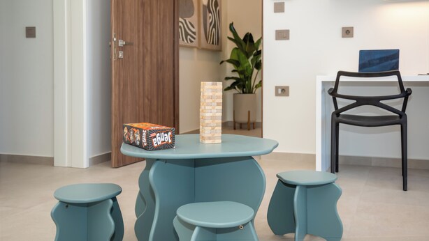 Una habitación moderna con una mesa color turquesa, dos taburetes a juego, una puerta de madera, una planta y un estante montado en la pared con cajas etiquetadas.