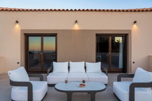 A modern outdoor seating area with a sofa and chairs, a table with drinks, and large sliding glass doors. - VaMa Luxury Living-8BR Design Villa with Heated Pool and Playroom (Episkopi Rethymnou)