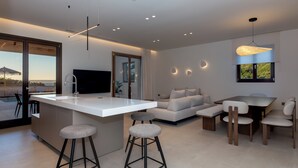 Una cocina moderna con una isla central, taburetes de bar, una zona de comedor con mesa y sillas, un sofá y un televisor montado en la pared.