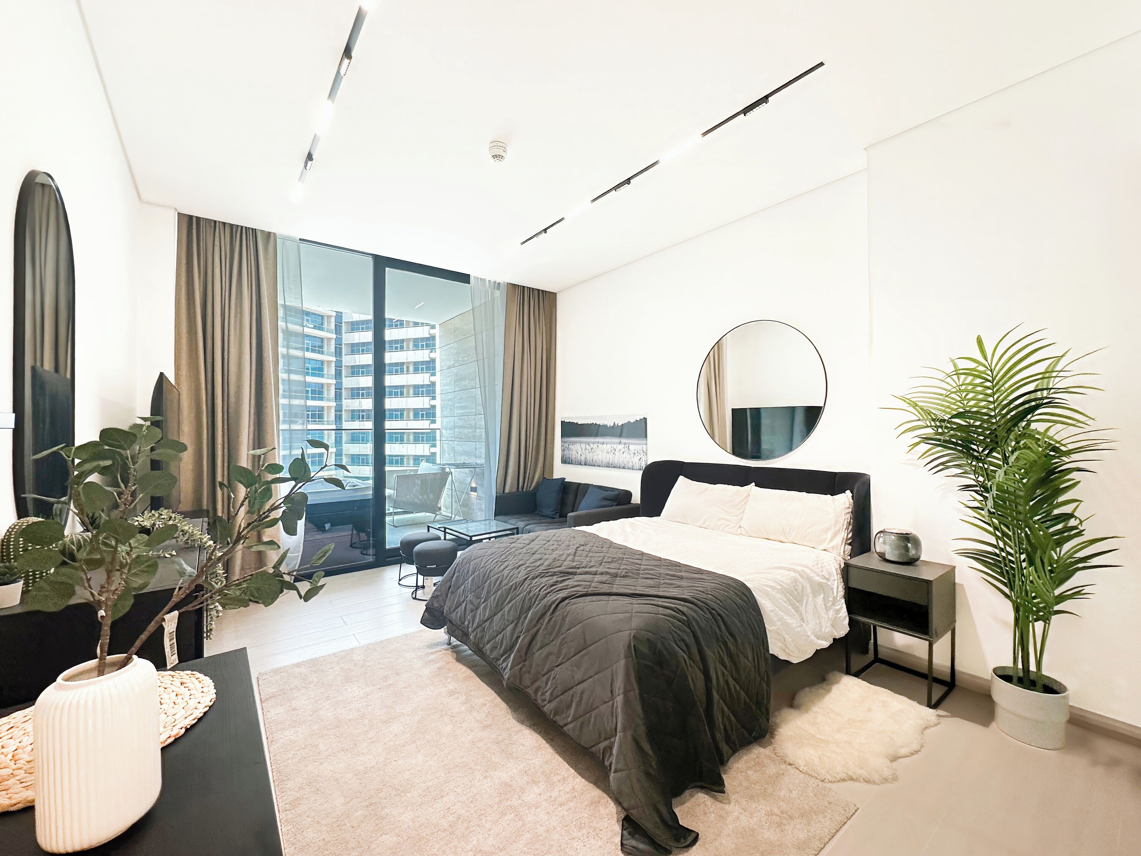 A modern bedroom with a large bed, a desk, a chair, a potted plant, and a view of a building through a large window.