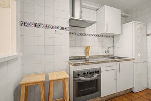 Una cocina con gabinetes blancos, estufa de acero inoxidable, horno empotrado, un lavamanos y dos taburetes de madera.