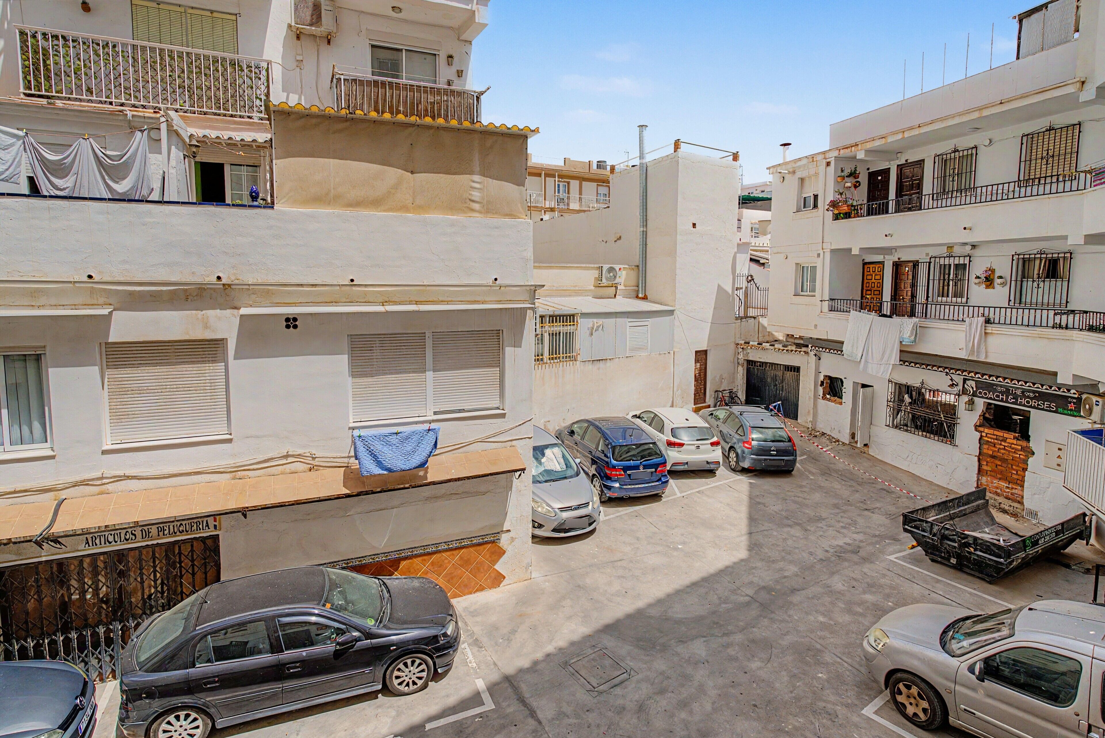 Una calle residencial con autos estacionados, balcones y una tienda con un letrero.