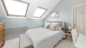 Um quarto com teto inclinado, claraboias, uma cama com almofadas, uma mesinha de cabeceira, um radiador, uma cômoda de madeira e uma porta.