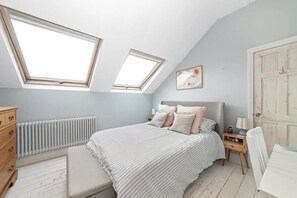 Ein Schlafzimmer mit schrägen Wänden, Dachfenstern, einem Bett mit Kissen, einem Nachttisch, einem Heizkörper, einer Holzkommode und einer Tür.