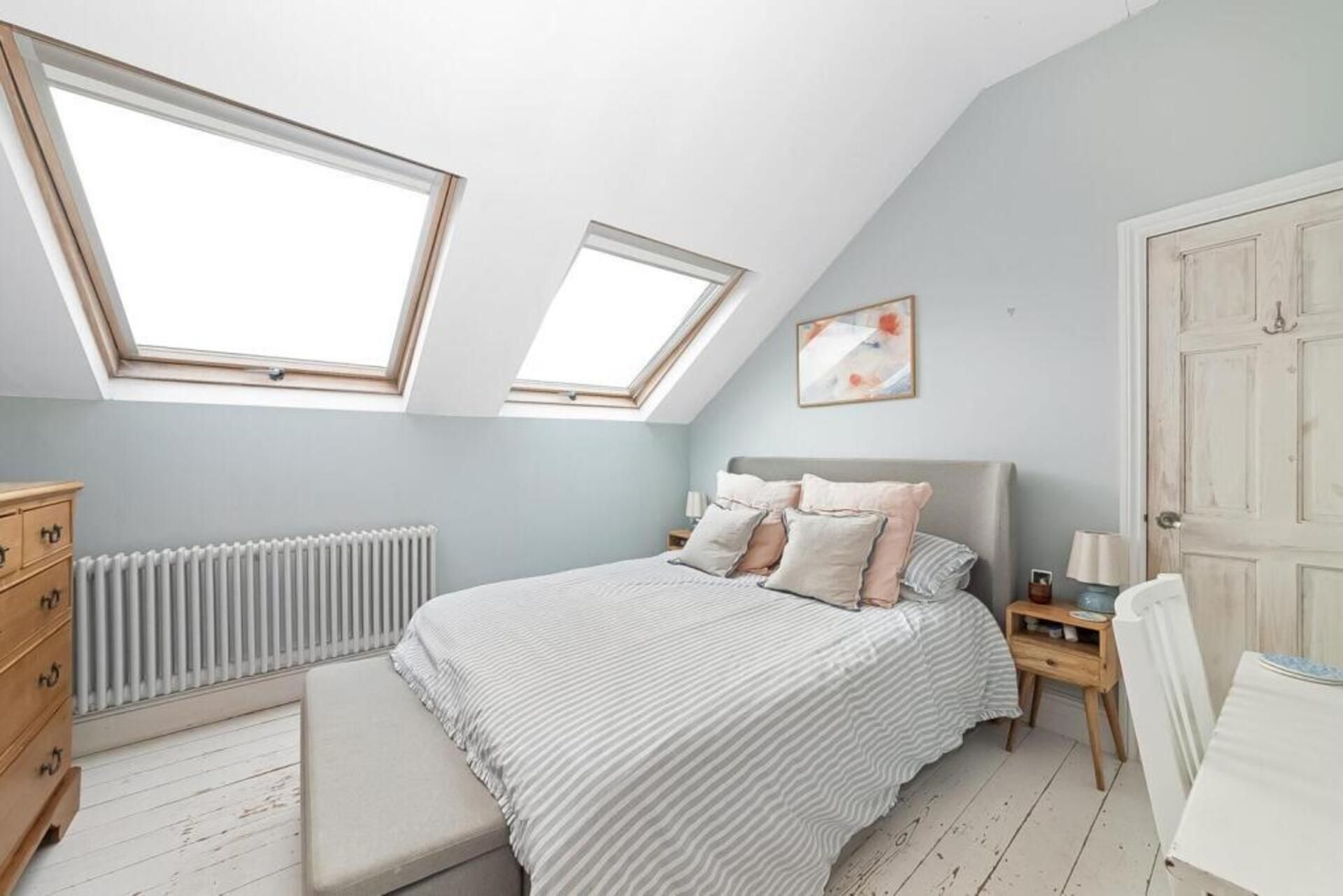 A bedroom with a sloped ceiling, skylights, a bed with pillows, a bedside table, a radiator, a wooden dresser, and a door.