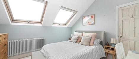 A bedroom with a sloped ceiling, skylights, a bed with pillows, a bedside table, a radiator, a wooden dresser, and a door.