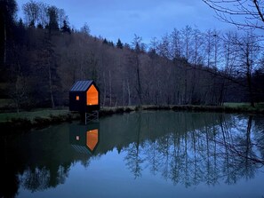 Une petite péniche de couleur sombre, dont l’intérieur est éclairé, flottant sur une étendue d’eau calme.