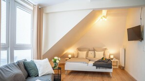 Ein Schlafzimmer mit schrägem Dach, einem Bett mit Kissen, einem Nachttisch mit Lampe, einem an der Wand montierten Fernseher und einem Fenster mit Jalousien.