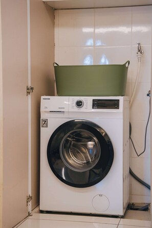 Eine weiße Waschmaschine mit einem grünen Deckel, platziert in einem Raum mit weißen Fliesenwänden und einer Tür.