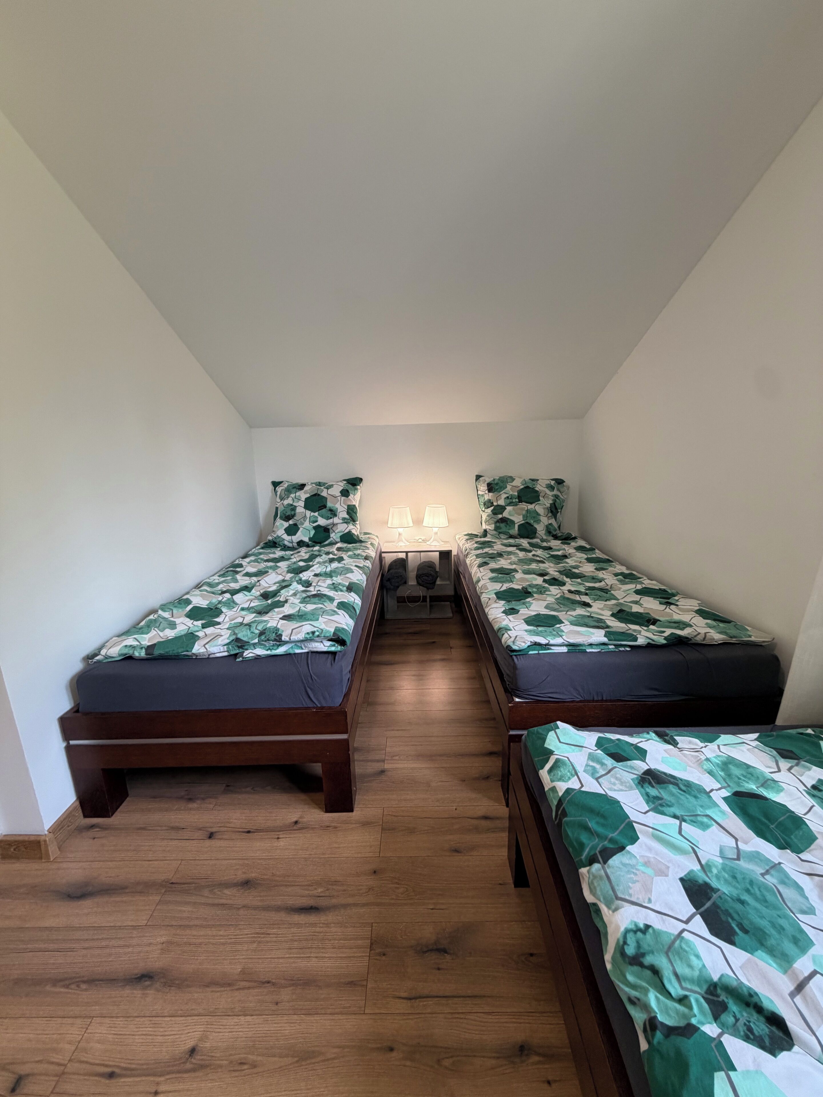 Pequeña habitación estrecha con dos camas de madera, cada una con colcha estampada. Dos lámparas de noche sobre una mesita entre las camas. El suelo es de madera y las paredes son blancas.