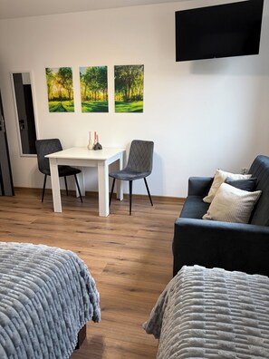 Una habitación moderna con una mesa blanca, dos sillas, un sofá y un televisor fijado a la pared.